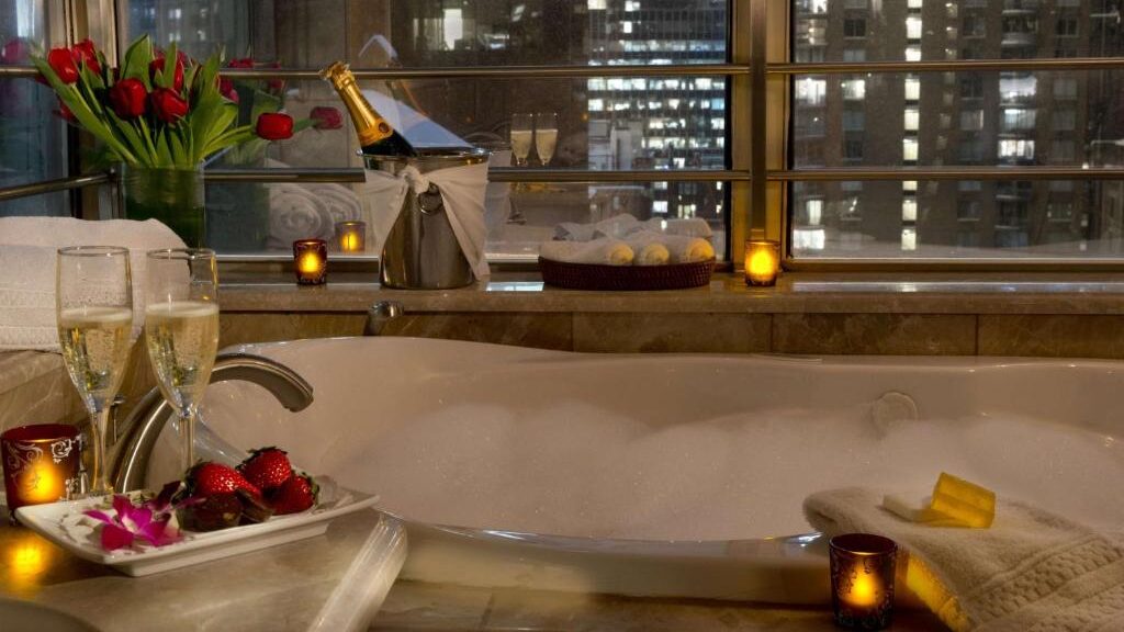 Romantic hotel bathroom with large soaking tub, champagne, candles, strawberries, and city night views